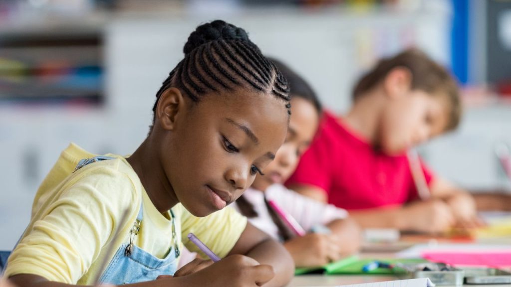 Three young students write in notebooks in a classroom. The girl closest to the camera is Black with braids pulled into a bun. Next to her are a girl and boy, each with brown skin.
