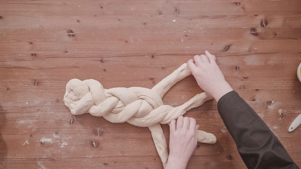 A pair of hands braiding challah dough on a flour-dusted wooden table.
