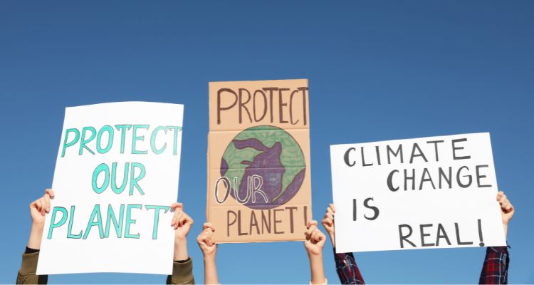Three pairs of light-skinned hands hold up cardboard signs with conservationist messages such as "Protect Our Planet!" against a clear blue sky.