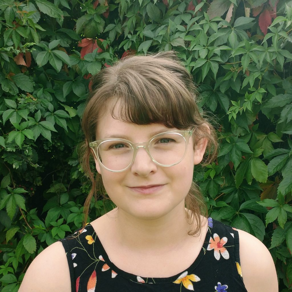 Rose Robbins, a smiling light-skinned woman with straight brown hair tied back and green eyes, wearing a flowered tank top and clear glasses, and standing in front of a wall of ivy.
