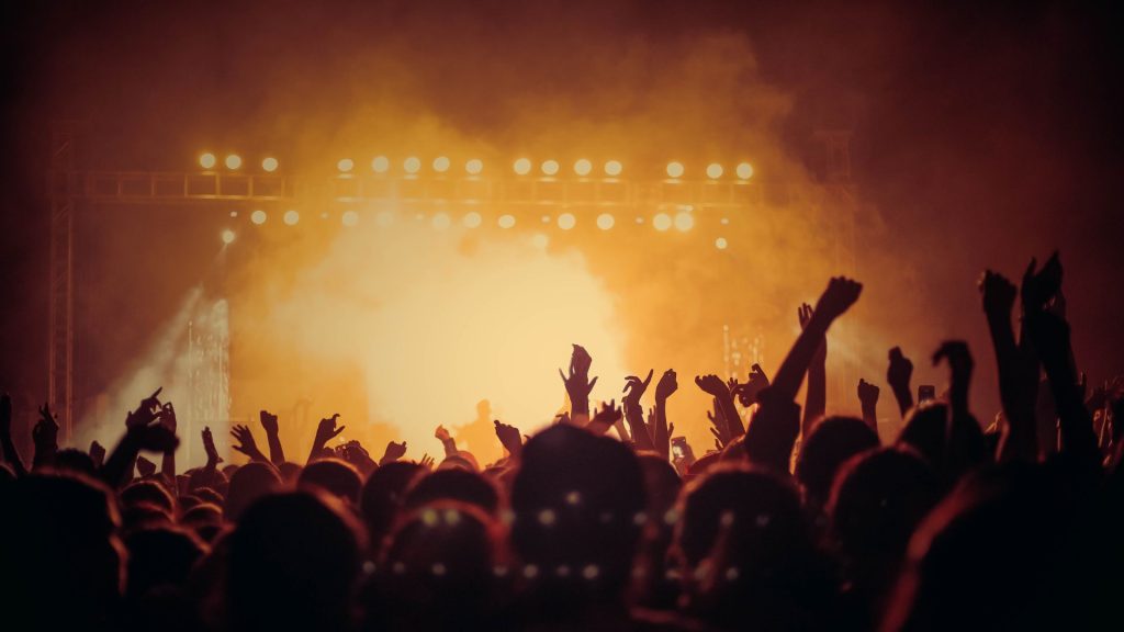 A crowd of concertgoers shot from behind, their hands raised. In front of them, a stage glows with smoke-diffused golden light against a dark night, and the silhouettes of band members can be seen.