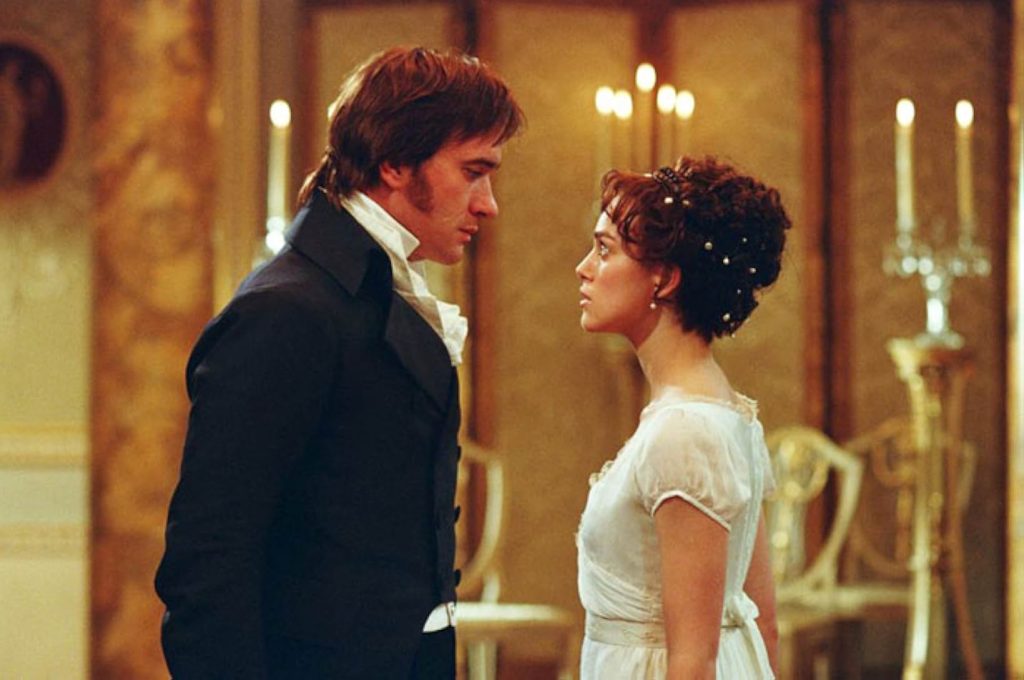 Mr. Darcy and Lizzie Bennet face each other in an empty ballroom with lit candles in the background. Mr. Darcy wears a black coat and Lizzie wears a white dress with pearls in her hair.