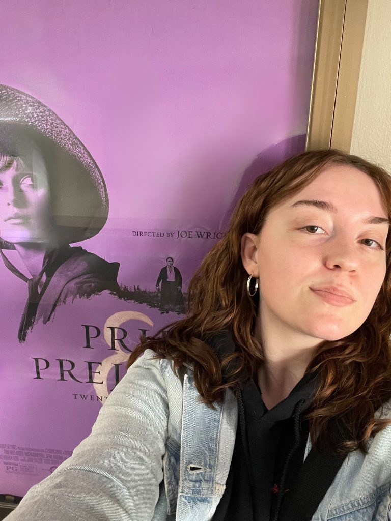 Kaija Strong wears a black hoodie, a blue denim jacket, and silver hoop earrings and stands in front of the Pride and Prejudice 20th anniversary poster. The poster is purple and features Kiera Knightley wearing a bonnet and looking to the side and Matthew Macfadyen walking through a grassy meadow in a long coat and open white shirt.