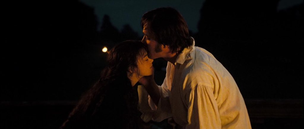Mr. Darcy wears a white shirt and kisses Lizzie Bennet on the forehead. She wears a brown jacket and closes her eyes while smiling softly. They are sitting outside at night.
