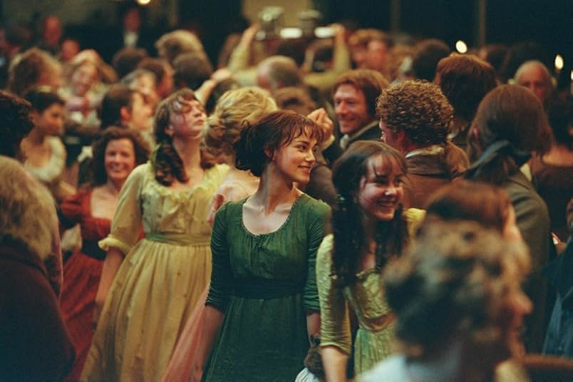A crowded ballroom full of smiling dancers. Prominently featured are Kitty Bennet in a yellow dress, Lizzie Bennet in a dark green dress, and Lydia Bennet in a cream dress.