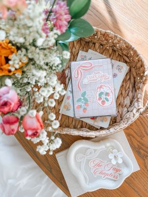 A basket with a stack of white, pastel-accented notepads sits on a wood floor. On the top of the stack is a sticker set with a sticker depicting a pink ribbon, a sticker reading "Cozy Girl Social Club", and two stickers depicting bunches of strawberries. To the left of the basket is a bouquet of flowers along with a white sheet. Below the basket is a small white dish sitting atop a white book. Inside the dish are a sticker depicting a pink ribbon, a sticker reading "The Faye Chapters" in pink cursive, and a white flower decoration.