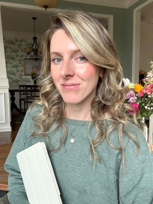 Kat of @thefayechapters, a light-skinned woman with long, wavy blonde hair, smiles closed-mouth at the viewer. She stands in three-quarter view facing the viewer's left, wearing a green sweatshirt and gold necklace, and holds a tablet in a white case. Behind her is a vase of flowers and the entry to a dining room in the background.
