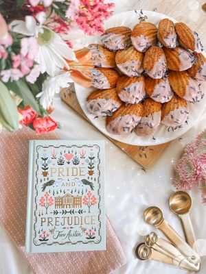 A flatlay displaying a copy of Jane Austen's Pride and Prejudice atop a pink napkin, below a white plate of pink-glazed madeleines atop a wooden board. Next to the book on the white table spread is a set of gold measuring spoons. Bracketing the display are pink, red, and white flowers.