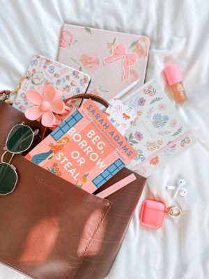A flatlay with three pastel notebooks and a copy of Beg, Borrow, or Steal by Sarah Adams spilling out of a brown leather bag, laid atop a white sheet. Also included in the display are a pink flower decoration, a sticker depicting a pink bow, a white stylus, a small tube of lip gloss, a pair of white AirPods and their pink case, and a pair of sunglasses.