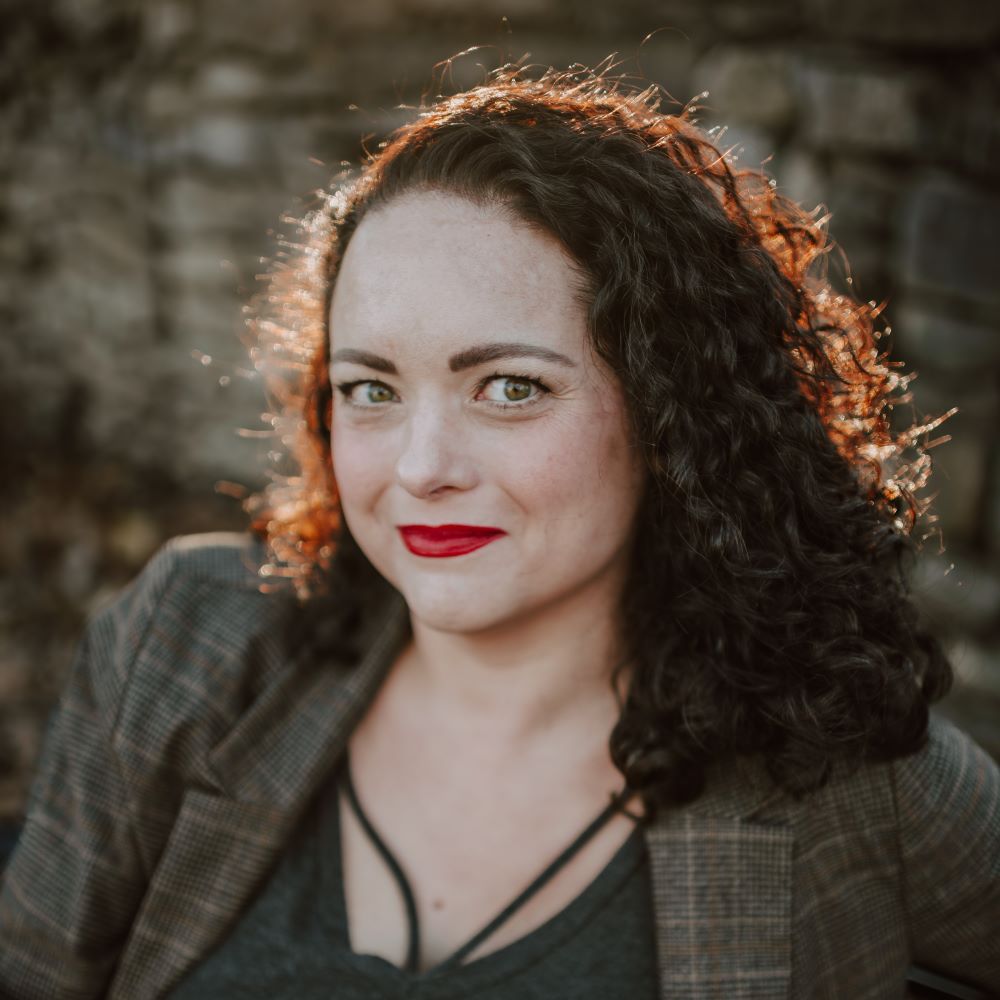 Cara Broel, a light-skinned woman with dark, curly shoulder-length hair and green eyes wearing read lipstick and a tweed blazer over a gray sweater.