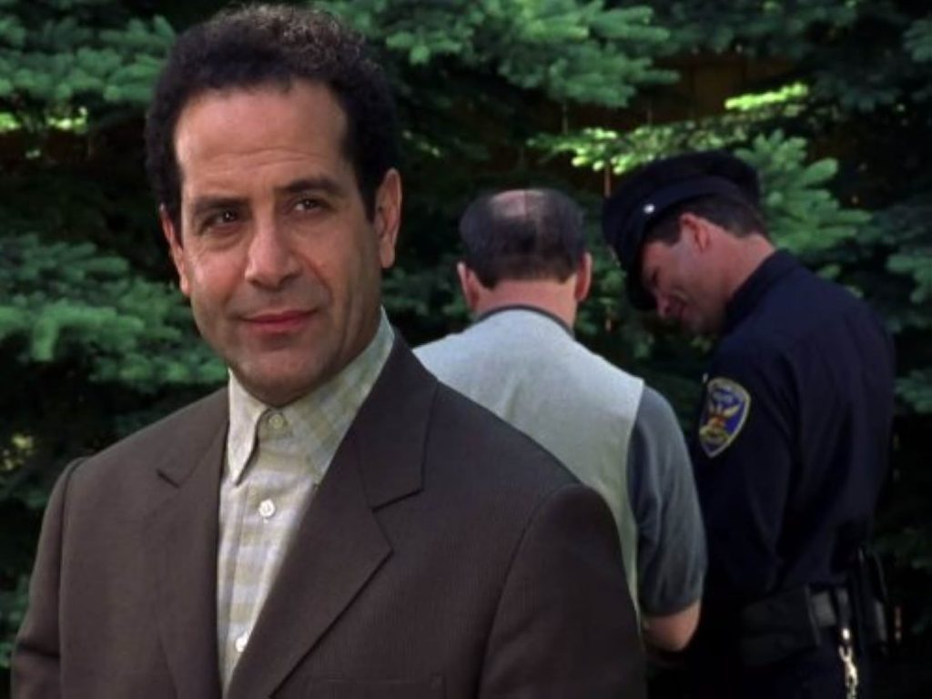 A man wears a brown suit and yellow plaid shirt. Behind him is a police officer and a man in gray shirt and white vest standing in front of pine trees.