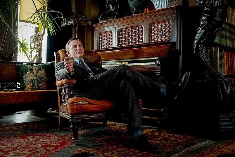 A man in a gray suit holds a coin in his hand and casually leans back in an orange antique armchair. Behind him is a wooden piano, a carved wooden statue, a matching antique sofa with a green and yellow pillow, a potted plant, and a fake skeleton. On the floor is a red and yellow rug.