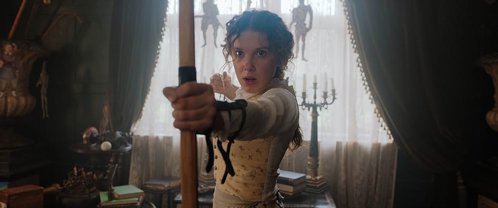 A woman in an old fashioned white blouse and yellow floral vest prepares to fire a bow and arrow. She stands in front of a desk full of books and candles and a window with white and green curtains.