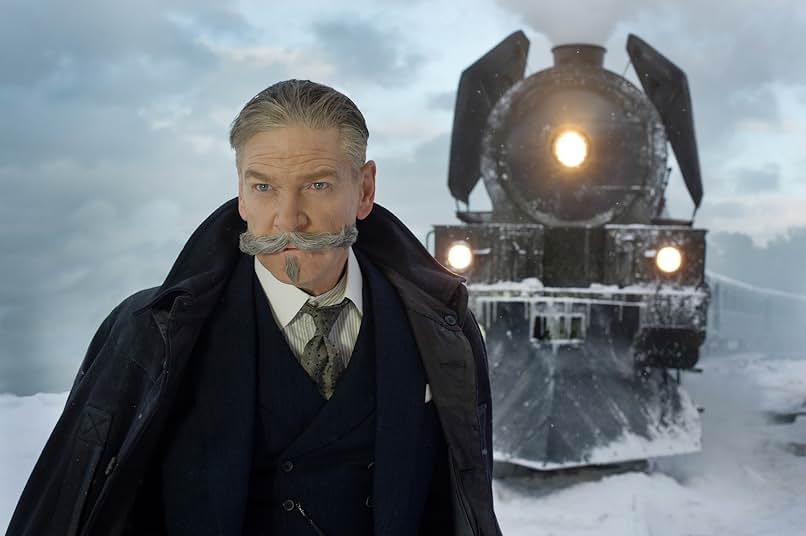A man with an old-fashioned gray moustache and pointed goatee wears a black suit, gray tie, and black coat. He stands in front of a train in the snow.