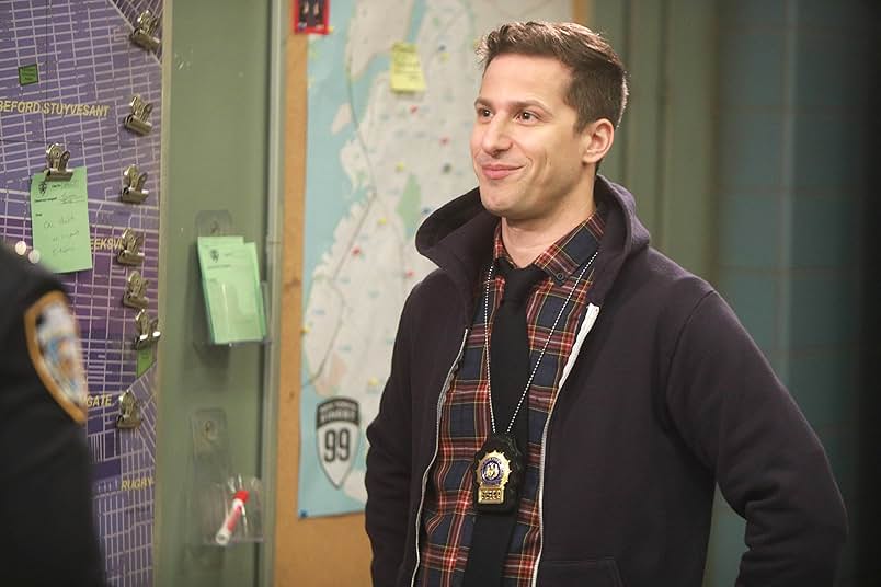 A man wears a black hoodie, a red and blue plaid shirt, a black tie, and a police badge on a chain around his neck. Behind him is a map covered with pins and pasted notes.
