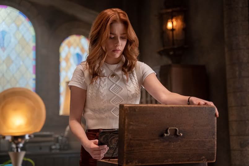 A redheaded girl wears a short sleeve white sweater and a gold necklace. She looks inside an opened antique wooden box. Beside her is a yellow globed lamp. Behind her are arched stained glass windows in yellow, blue, pink, and green.