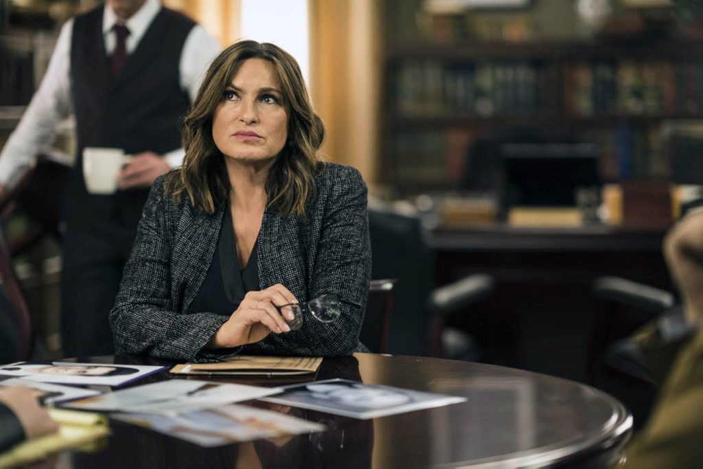 A woman in a dark gray suit and black blouse sits at a round table and holds a pair of glasses in her hand. Scattered on the table are criminal files. Behind the woman is an office with a walled bookshelf and a man in a black vest holding a white mug.
