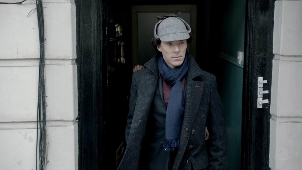 A man with a gray hat, black coat, black suit, maroon shirt, and blue scarf steps through an open doorway. Beside him are white stone walls. The wall of the entrance hall behind him is dark green, and the door is black.