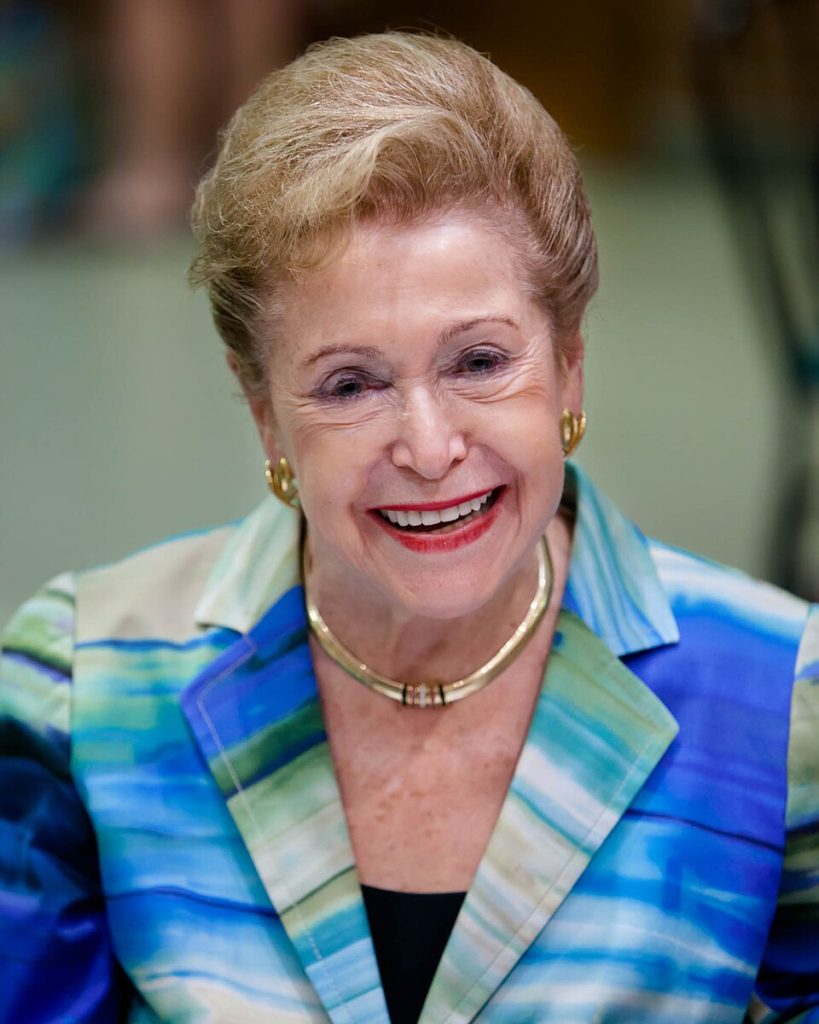 Mary Higgins Clark smiles and wears gold earrings, a gold necklace, and a blue and green abstractly striped suit. Behind her is a green blurred background.