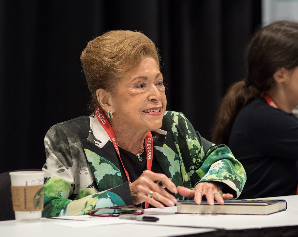 Mary Higgins Clark smiles as she signs books at a table. She wears gold and green earrings with a matching necklace, a green, black, and white suit, a black top, and a red lanyard with the name BookBub printed on it in white lettering. Behind her is a black curtain.