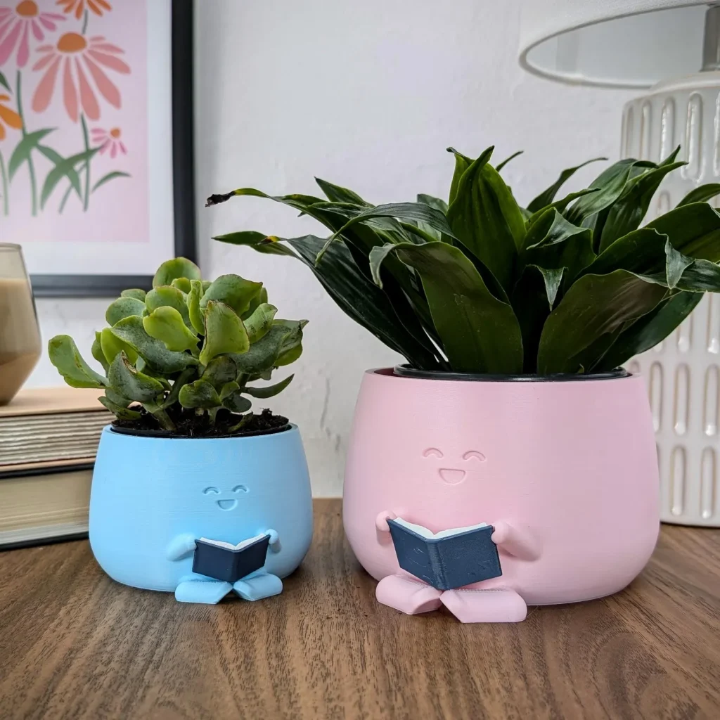 Two planters are sitting on top of a brown table. The planters have smiley faces in the center, two little legs, and two little arms holding a small ceramic book. The planter on the left is light blue and smaller than the light pink planter on the right. Each planter has leaves growing out of the top, making it look like the reading planter characters have hair.