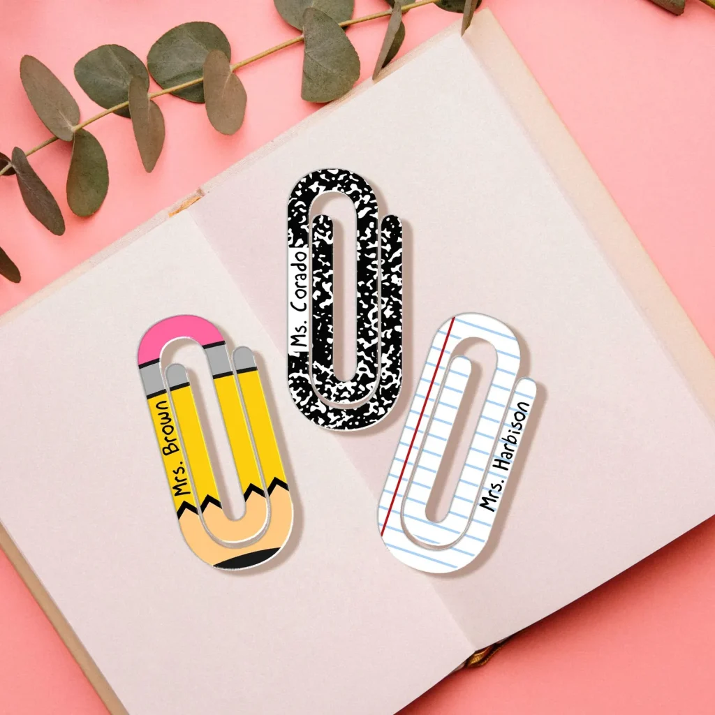Three acrylic paperclip-styled bookmarks. One paper clip has the outlines and colors of a pencil, the other one has the same colors and patterns as a composition notebook, and the last one is modeled after binder paper. All three bookmark paper clips have a teachers name on the side.