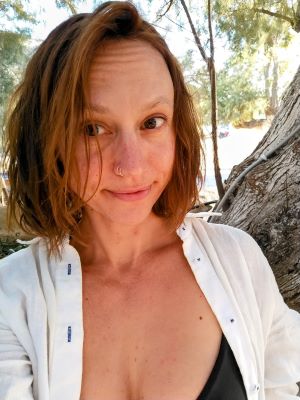 Image of Trish Fancher. Trish has short brown hair and a nose piercing and looks at the viewer with a closed-mouth smile. She is wearing a white sweater and black top and is in front of a tree with water and more trees in the background. The sky is clear and sunny.