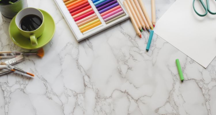 A cup of coffee, pastels, colored pencils, and other art supplies on a white marble surface.