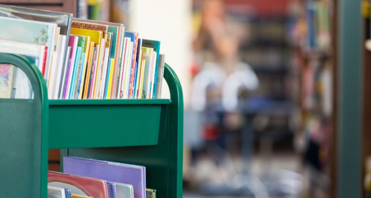 A green library cart filled with picture books on the top and bottom shelves within a larger library or bookstore.