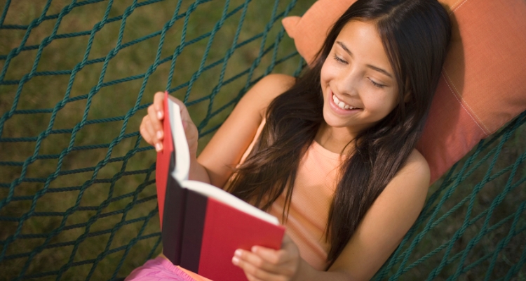 A girl is smiling reading a red book while she lays on a pillow in a hammock outside.