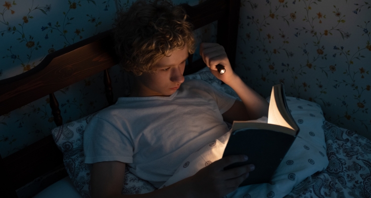 A boy is holding up a flashlight while he reads a book in his bed at night.