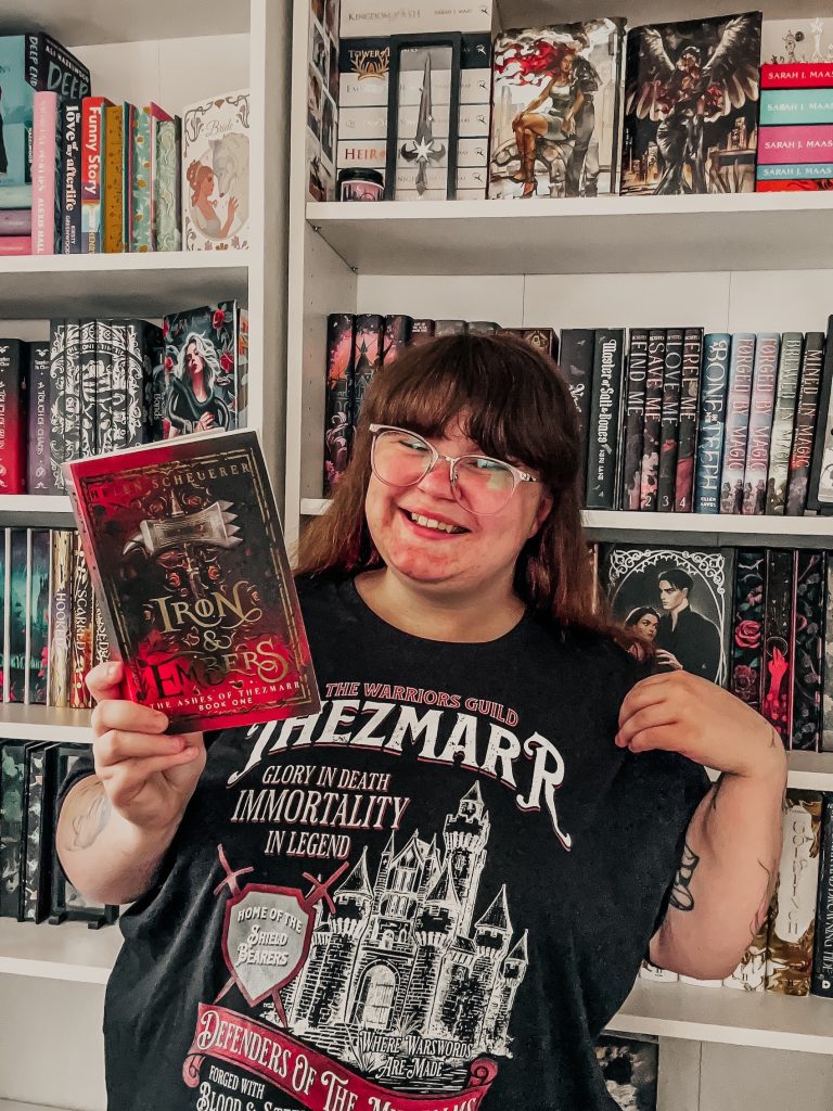 Danielle wears glasses and a black Thezmarr t-shirt while holding a copy of the book Iron & Embers. She smiles in front of a white bookshelf full of romance novels.