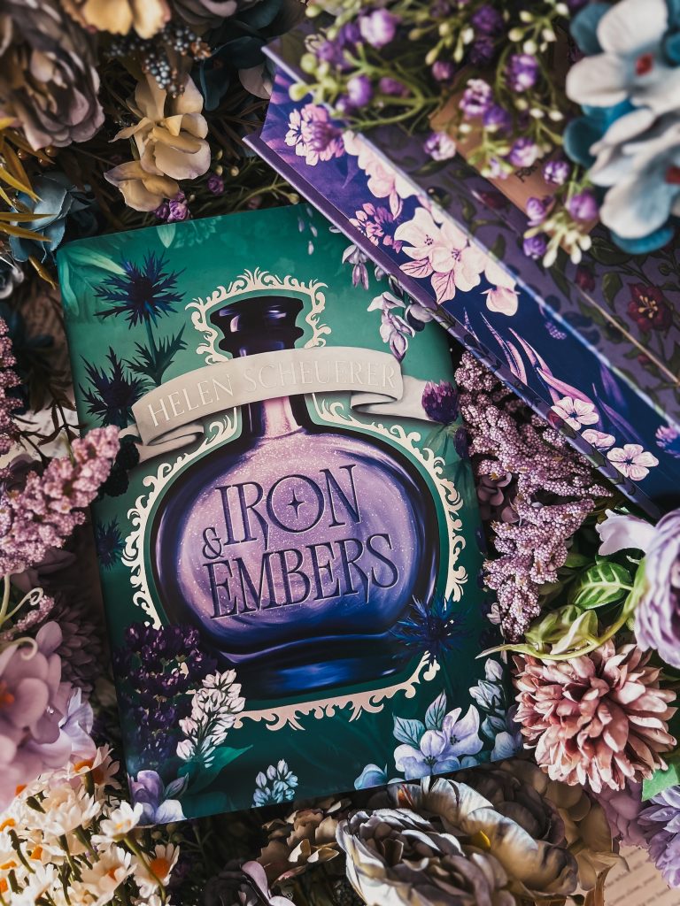 The copy of the book Iron & Embers by Helen Scheuerer is surround by purple, pink, and yellow flowers. The book cover has a teal background with a purple potion surrounded in a white swirling border. There are purple flowers around the potion. In the upper right corner is a purple book with the page ends painted purple with pink flowers.