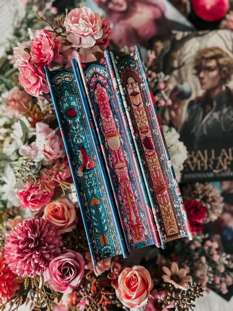 Three books with page ends painted like stained glass windows. The one on the left is mostly blue with a red apple, orange fox, and golden arrow. The center book is pink and blue with a gold goblet. The one on the right is red and yellow with a red anatomically correct heart and a golden crown. The books are nested in a pile of pink and white flowers. In the blurred background is another book cover with a brooding blond man in armor on the front.