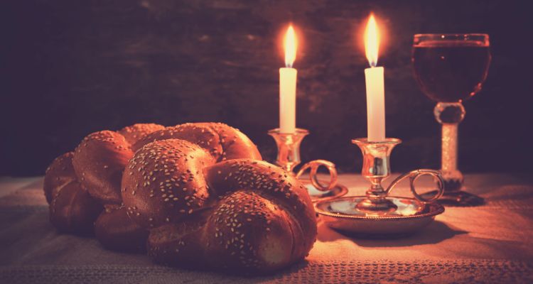 A loaf of challah bread, two taper candles in candleholders, and a glass of red wine sit on a table in a dimly lit space.