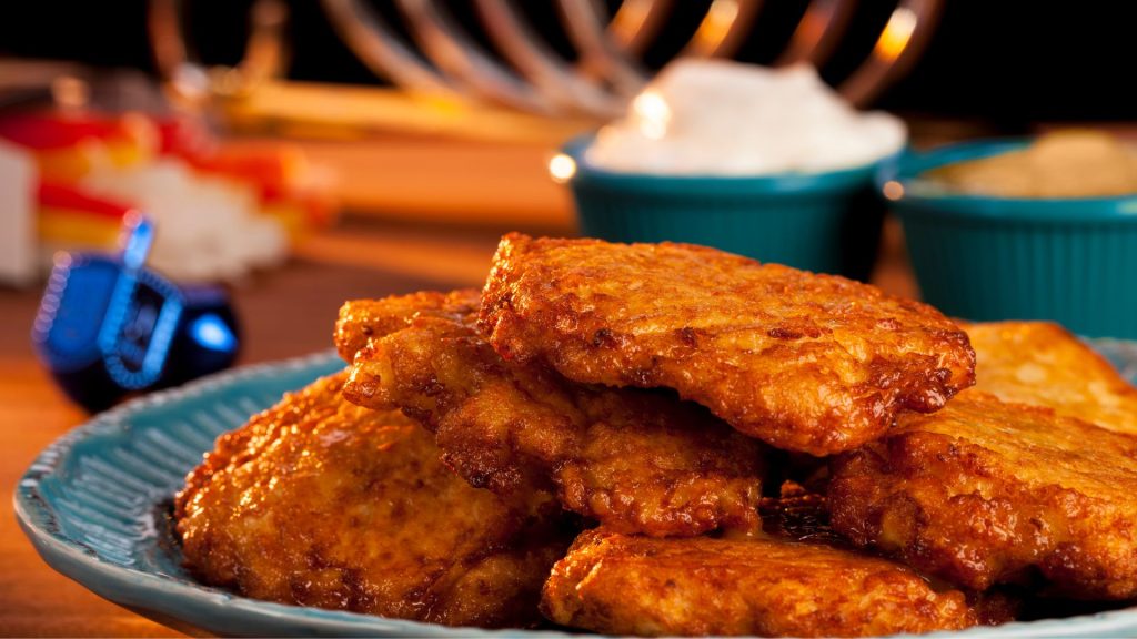 A plate of latkes on a warmly lit table. Out of focus in the background are two blue dreidels, small bowls of sour cream and applesauce, and a menorah.
