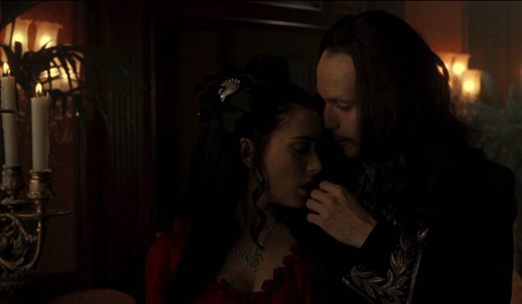 A still from the movie Bram Stoker's Dracula. A woman in a red Victorian dress with a silver hair comb turns passionately towards a man in a dark jacket trimmed with silver embroidery. He gazes down at her with adoration. Behind them is a dimly lit room with red walls and lit sconces.