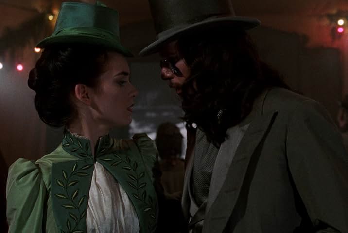A still from the movie Bram Stoker's Dracula. A woman in a green Victorian jacket and top hat with a white blouse faces a man in a gray Victorian suit and top hat. The man wears tinted glasses at the tip of his nose. They stand in a room with red walls in front of an open doorway.