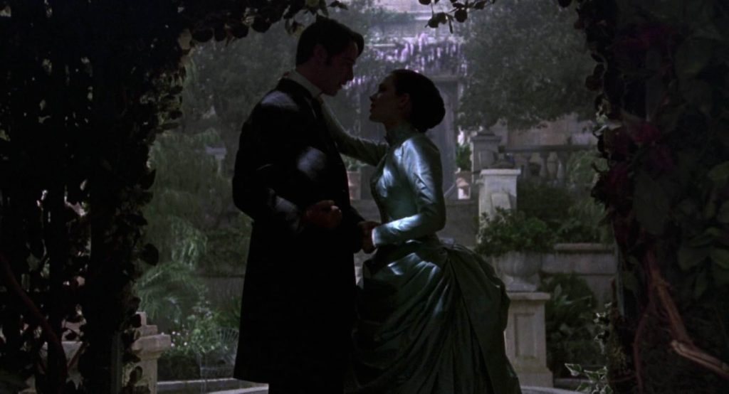 A still from the movie Bram Stoker's Dracula. A man in a long coat holds hands with a woman in a light green Victorian gown. They stand in an ornate garden and are framed by the shadow of a vine-covered bowery.