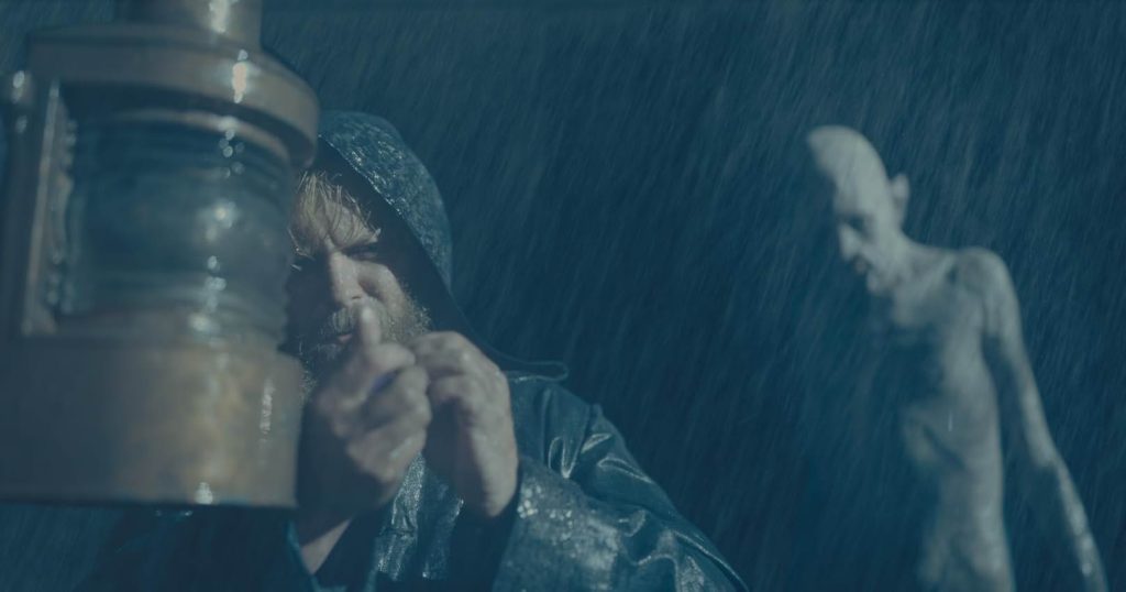 A still from the movie The Last Voyage of the Demeter. A man in a black raincoat stands against the curved wood of a ship's bannister in a rainstorm. Behind him is the pale, naked figure of a monstrous vampire.