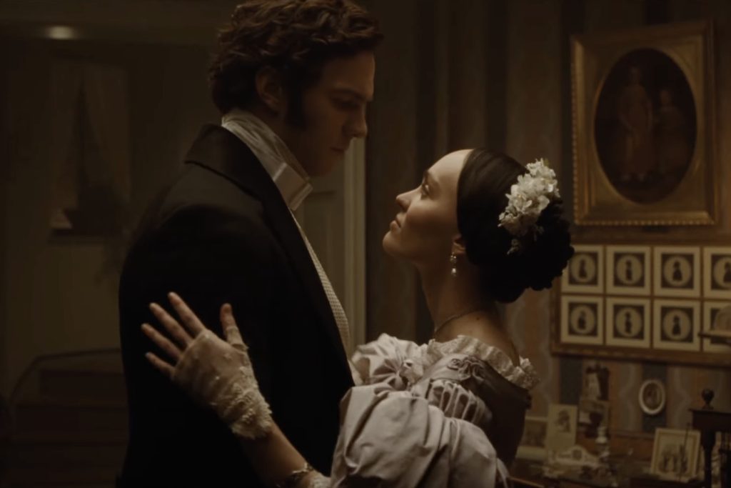 A still from the 2024 movie Nosferatu. A man in a dark coat embraces a woman in a purple Victorian dress. She wears white flowers in her hair and white lace fingerless gloves. Behind them is a brown papered wall with framed cameo pictures.