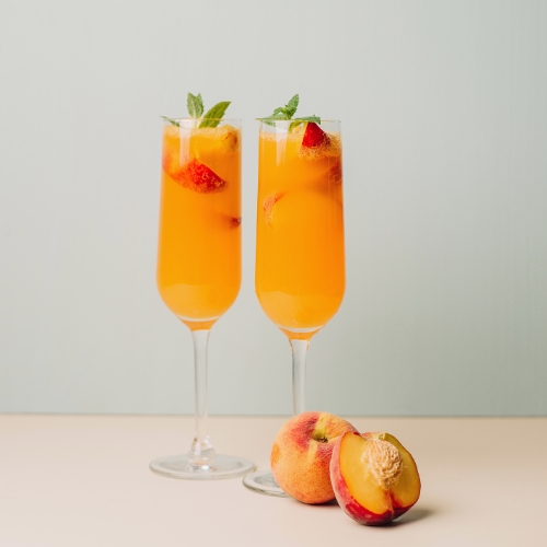 Two bright orange belinis in tall champagne flutes with a peach and mint garnish and a halved peach on the table next to it.