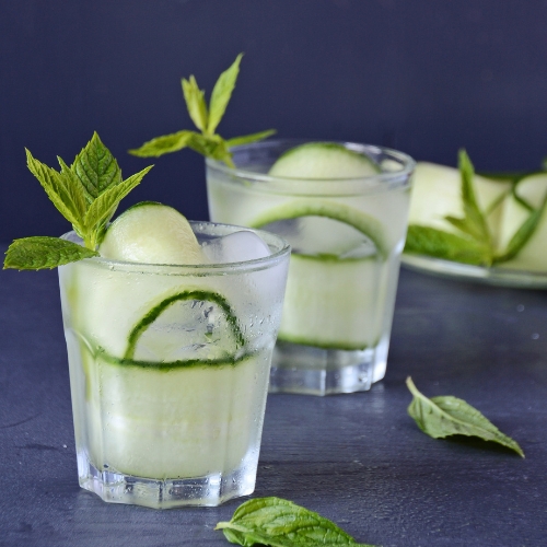 A cloudy light green drink sits in a ridged frosted glass with cucumber and mint leaf garnishes on a black table and surrounded by mint leaves.