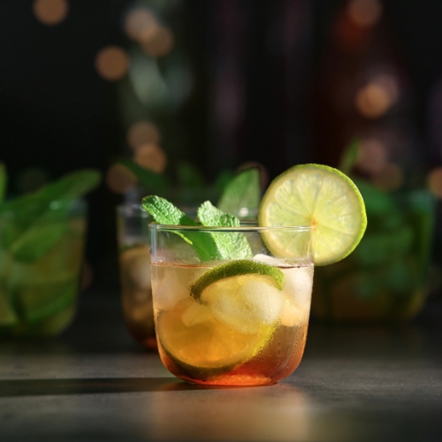 A light brown mint julep with ice, lime, and mint sits on a black surface.