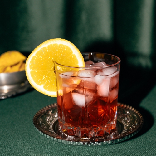 A dark red-brown liquid in an etched crystal glass with ice and a lemon wedge sits on a fancy metal coaster on a green table.