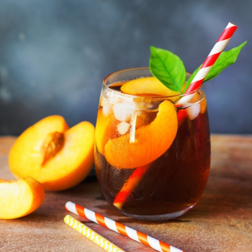 A dark brown peach iced tea is in a stemless wine glass with a leaf garnish and a red and white straw.