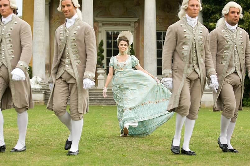 A still from Austenland. A woman in a light blue Regency gown, bonnet, and white lace gloves walks behind four men in tan Regency coats and powdered wigs. Behind them is a regal building with columns and topiaries.