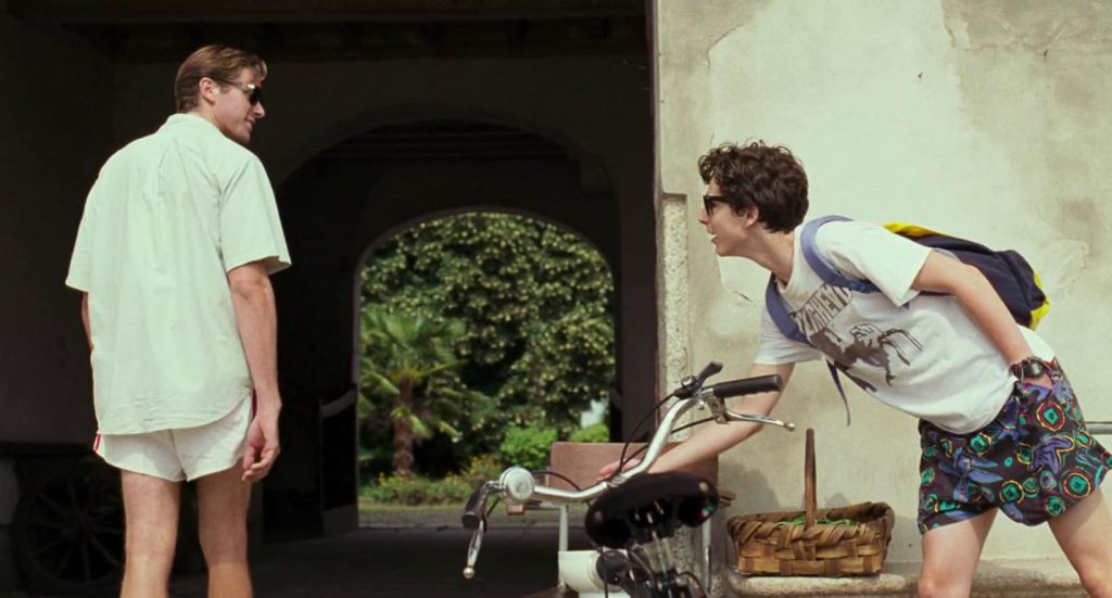 A still from Call Me By Your Name. A young blond man in a white buttoned shirt, white shorts, and sunglasses walks into stone tunnel leading to trees. He looks over his shoulder at another young man with curly brown hair wearing a white graphic t-shirt, black, blue, and red patterned shorts, and blue and yellow backpack. The brunette man wears sunglasses and leans over a resting bicycle. A basket is behind him.