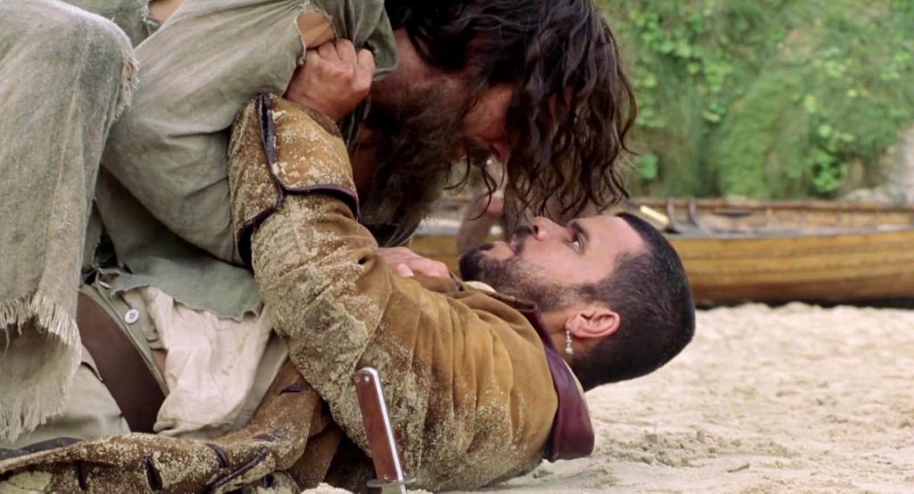 A still from The Count of Monte Cristo (2002). A man in gray tattered rags and wild hair grapples on the beach with another man in a tan, leather-trimmed coat wearing an earring.