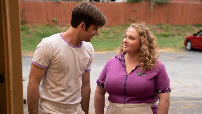 A still from Dumplin'. A young man and woman stand in a parking lot with a wooden fence. The man wears a white t-shirt with a purple collar and has a toothpick in his mouth. Looking at him is a woman wearing a purple waitress uniform and a black nametag that says Will.